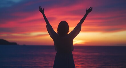Silhouette of a woman with raised arms against a sunset sky over a sea background.