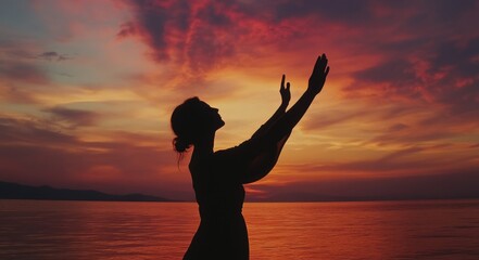 Fototapeta premium Silhouette of a woman with raised arms against a sunset sky over a sea background.