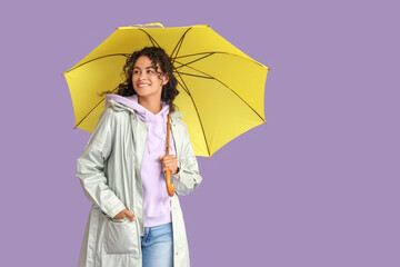 Happy young African-American woman in raincoat with umbrella on lilac background