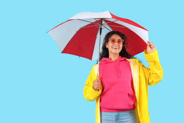 Happy young African-American woman in raincoat with umbrella on blue background