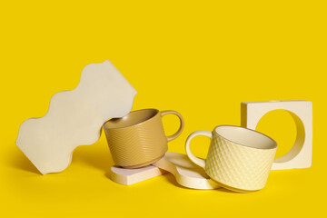 Different ceramic mugs and plaster podiums on yellow background