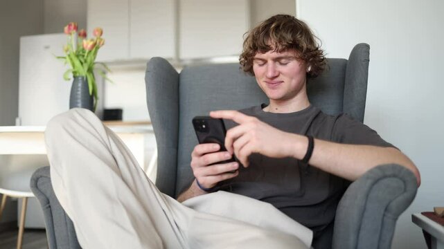 Happy and engaged, a young man sits in a chair, smiling as he interacts with his phone. His fingers are actively tapping the screen, fully enjoying whatever task or game he's immersed in.