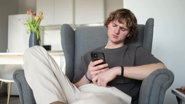 Very tired A young man sitting in an armchair with his head tilted back, holding his phone in his hand, looking exhausted and deeply tired, possibly after a long day or intense activity.