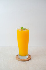 Tall Glass Of Fresh Mango Juice Garnished With Mint Leaves On A Wooden Coaster Against A Clean Minimalist Background.