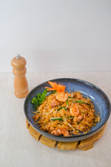 Stir-Fried Kwetiau Noodles With Shrimp And Vegetables Served In A Black Bowl With Carrot Garnish And Pepper Grinder On Light Background.