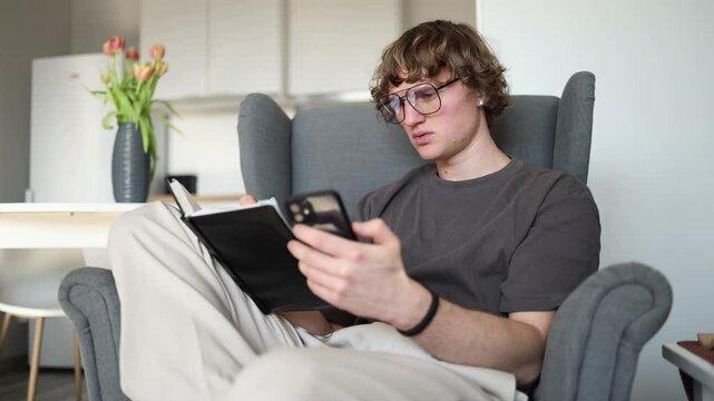 A young man sits in a chair, holding a phone in one hand and a notebook in the other, deep in thought, giving off a studious and focused vibe.