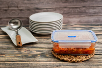 Glass container with food inside next to a stack of plates and a ladle, on a dark rustic wooden background, close-up.