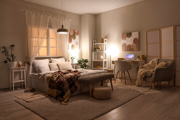 Interior of stylish cozy bedroom with laptop on workplace and glowing lamps in evening