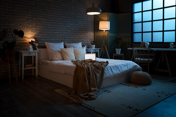 Interior of bedroom with laptop on bed and glowing lamps at night