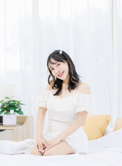 Asian happy young beautiful cheerful female sitting on cozy white bed smiling looking around in bedroom at home, Good morning calm comfortable healthcare relaxing resting well being peaceful concept.