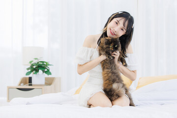 Asian happy young female sitting on white bed owner smiling holding playing fluffy tortoiseshell cute little kitten breed pussycat pet friend in bedroom at home, Concept love animal comfort rest care.