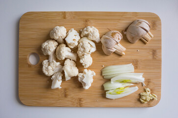 Close-up of a bamboo cutting board with chopped cauliflower, mushrooms, onion, and garlic. Natural home lighting, top-down view on a white table. Perfect for cooking themes
