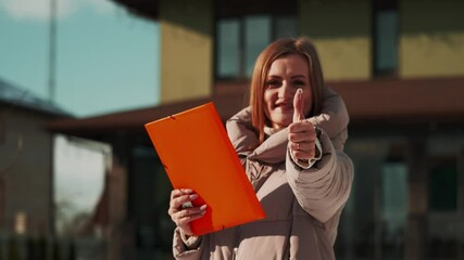 Professional realtor woman showing thumbs up gesture while holding orange folder with property documents, standing outdoors near residential building