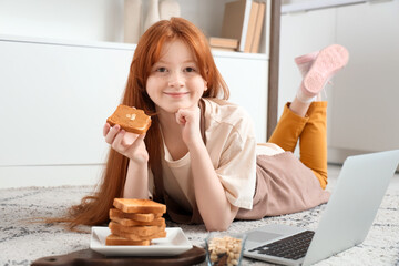Teenage redhead girl eating toast with nut butter and laptop on floor in kitchen