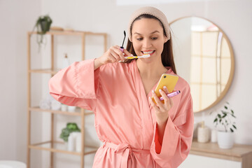 Hurrying young woman with mascara and mobile phone brushing teeth in bathroom
