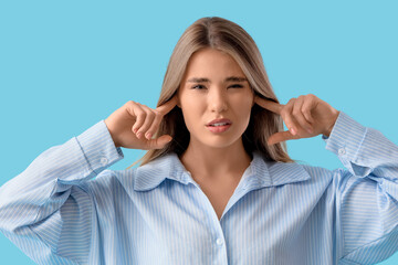 Young woman in pajamas suffering from loud noise on blue background, closeup