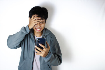 Asian teenager in white t-shirt and blue hoodie, posing angrily and covering his face while looking at his cellphone