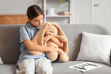 Sad girl with toy bunny and family figure sitting on sofa at home. Adoption concept