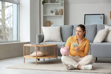 Fototapeta premium Young woman with piggy bank and dollar banknotes sitting on floor at home