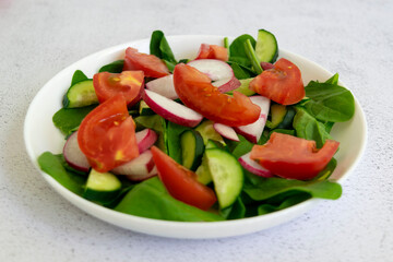 Raw fresh green healthy organic salad of spinach, cucumber, tomato and radish in white bowl on table top view on grey background.