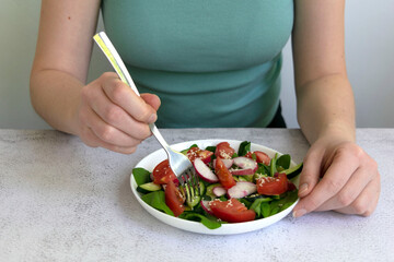 Woman hands cooking fresh healthy organic salad of spinach, cucumber, tomato and radish in white bowl on table top view on grey background.