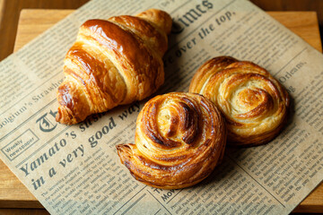 新聞紙風のオーブンペーパーに乗せた焼き立てクロワッサンとシュガーロールデニッシュ　木製テーブルの背景