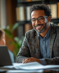 A man wearing glasses is smiling and sitting at a desk with a laptop and papers