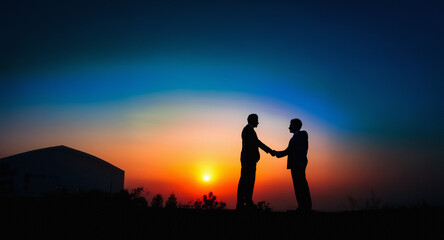Fototapeta premium Two people shake hands in silhouette against a vibrant sunset sky. A building silhouette stands to the side, creating a scene of connection and agreement against the colorful horizon.