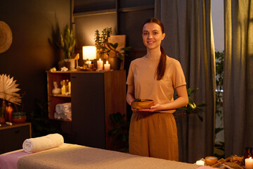 Portrait of young Caucasian masseuse with long ponytail dressed in beige uniform holding wooden...