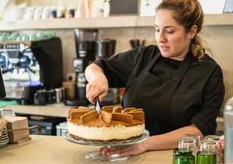 Woman barista cutting a caramel cheesecake with a knife in a small bakery business.