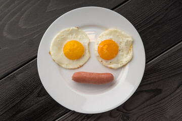 Two fried eggs with sunny side up and a single sausage on a white plate over a dark wooden background.