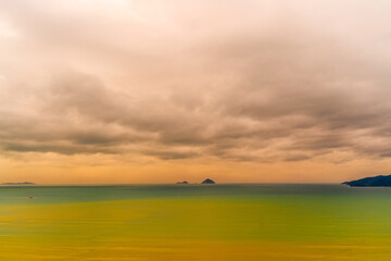 Fototapeta premium A serene view of the ocean featuring two distant islands. The sky is filled with soft, gray clouds, reflecting warm hues of sunset. Gentle waves lap against the shore