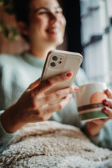 Young caucasian woman use phone send sms text messages and drink coffee by the window