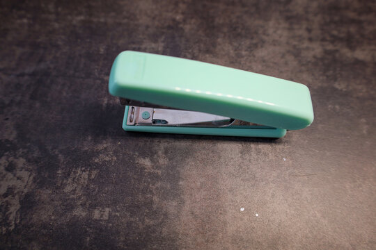 A small mint-colored office stapler is depicted on a dark gray background. The stapler is positioned at a slight angle, its top part is open. Staples for the stapler are not visible.