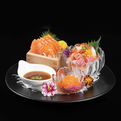 Fresh sashimi platter featuring salmon and assorted seafood garnished with flowers and sauces, beautifully presented on black background