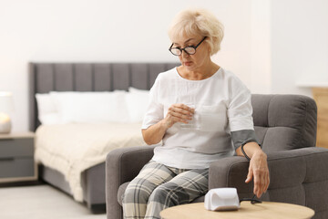 Senior woman measuring blood pressure in armchair at home. Space for text