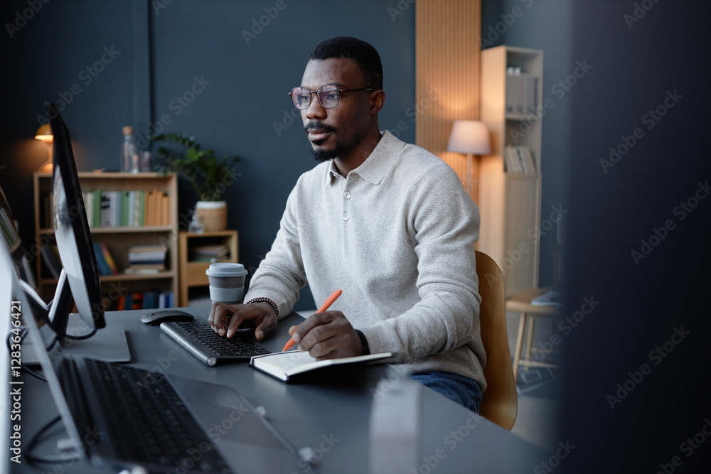 Poster focused black man with glasses looking at computer screen writing in notebook at working station in 
