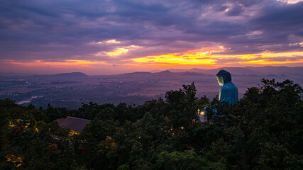 A serene aerial view of a large Buddha statue surrounded by lush forest, with a stunning sunrise...