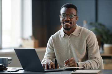 Portrait of cheerful adult Black man wearing glasses captured while writing in notebook at desk with laptop in cozy home office, copy space