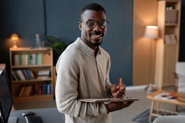 Portrait of adult Black man beaming with smile while looking at camera holding clipboard with notes working in modern office, copy space