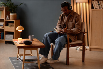 Full length shot of concentrated Black man in casual outfit listening to music through headphones, while writing in notes on paper sitting in vintage armchair in cozy office