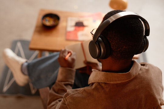 Rear view of Black man listening to podcast through wireless headphones while making notes in notebook in home office sitting at coffee table, copy space - Powered by Adobe