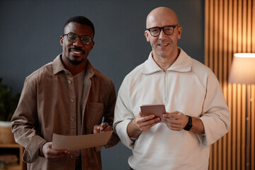 Waist up shot of bald mature male designer holding digital tablet posing next to Black male colleague cheerfully looking at camera, while drawing sketches on paper in office