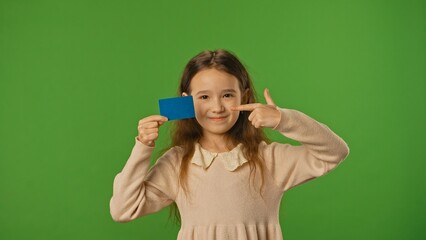 Little girl child holding advertisement card and pointing finger at blue space, positive expression, isolated on chroma key green screen background mockup.