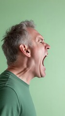 Fototapeta premium Profile view of a furious mature man yelling with his eyes closed and mouth wide open, expressing extreme anger and frustration against a vibrant green backdrop