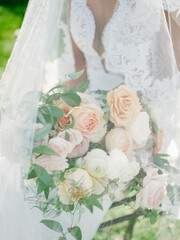 Bridal bouquet featuring pastel roses held by a bride in a white lace gown in a sunny outdoor...