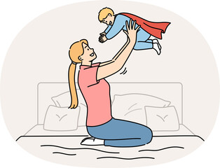Happy mother lifts up newborn son in superhero costume, sitting on bed in bedroom. Caring casual mother plays with child, motivating son to achieve ambitious goals and success in life