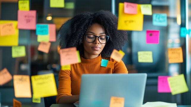 Young project manager using laptop and organizing work with colorful sticky notes on glass wall in modern office, focusing on efficient task management and deadline prioritization