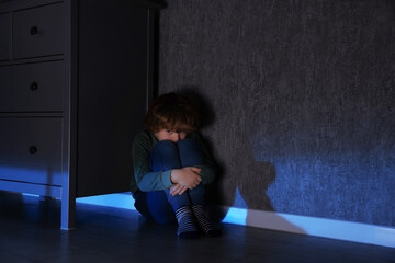 Scared boy hiding behind chest of drawers at night