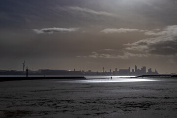 Fototapeta premium Lone person in silhouette walking dog on beach with city of Liverpool skyline in distance